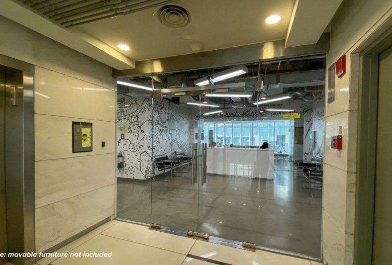 Modern office lobby with glass double doors, a white reception desk, and seating area beyond; mural wall behind.