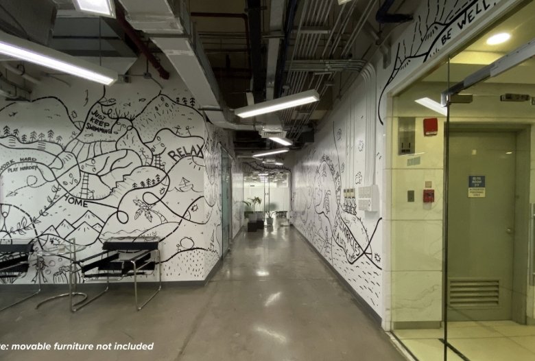 Long office hallway with white mural walls, exposed ceiling pipes, and hanging lights; left side has chairs, right has a glass door.