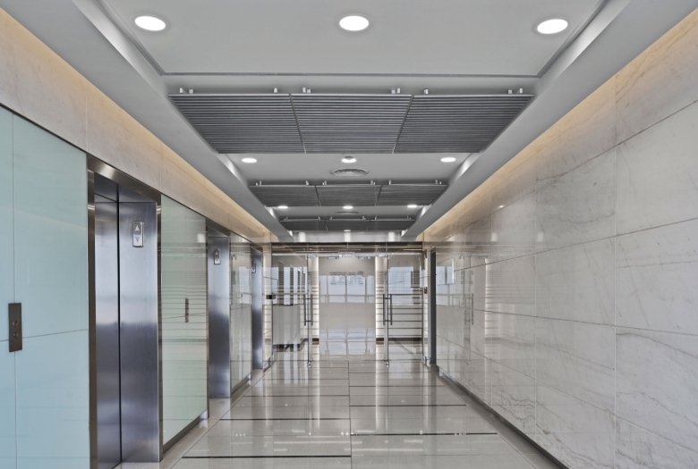 Empty modern office corridor with glass-walled offices on the left, two elevators, and a glass double door at the far end.