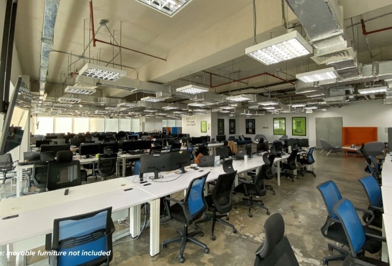 Open-plan office with long white desks arranged in rows, ergonomic black and blue chairs, and exposed ceiling ducts and lighting fixtures in a spacious workspace. Note: movable furniture not included.