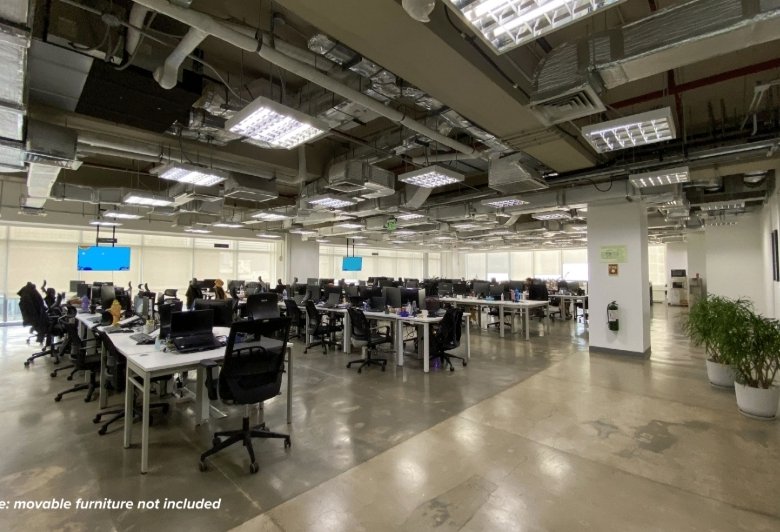 Open-plan office with rows of desks, computers, and black chairs under an exposed industrial ceiling.