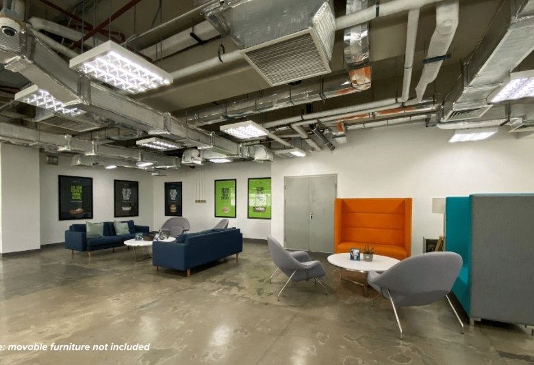 Industrial-style lounge with blue sofas, gray chairs, round white tables, green-framed posters, and exposed ceiling ducts. Note: movable furniture not included in the image.
