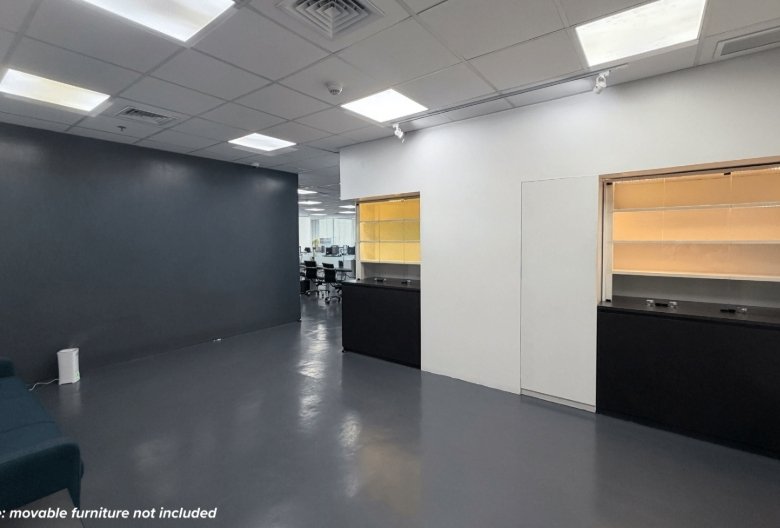 Vacant office showroom with gray floor, dark accent wall on the left, and two service counters with shelves on the right; teal seating visible in the corner.