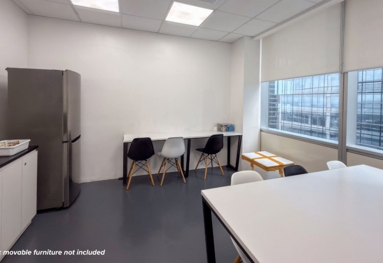 Office break room with a stainless steel fridge, white cabinets, and a long counter with four chairs by a large window.