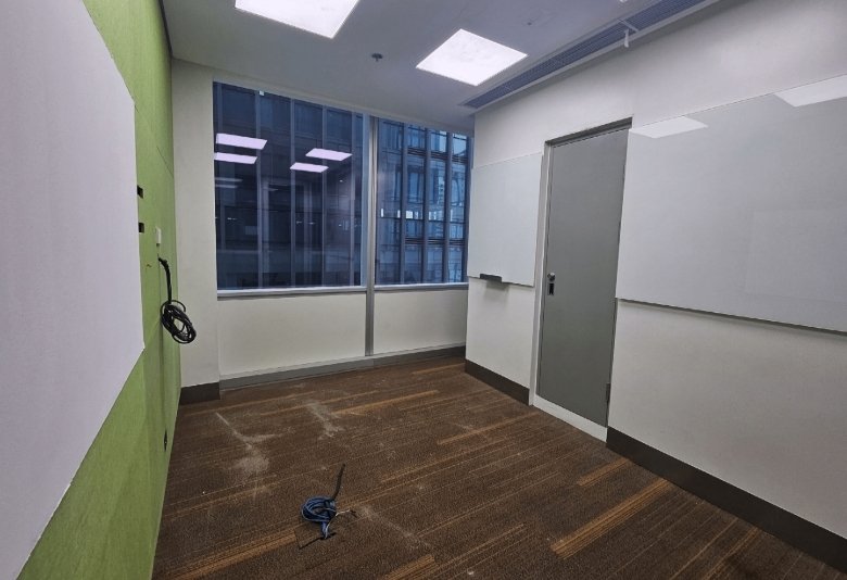 Empty office room with a green accent wall, whiteboards, large window, and a closed door; cables lie on a brown carpeted floor.