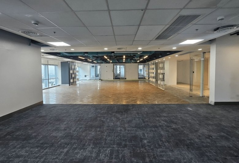 Empty office space with dark carpet in the foreground and an open area with a wood floor at the back, separated by glass partitions.