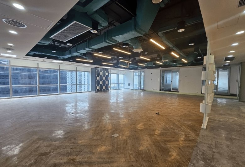 Empty modern office space with exposed ceiling ducts and large windows on one side, under renovation.