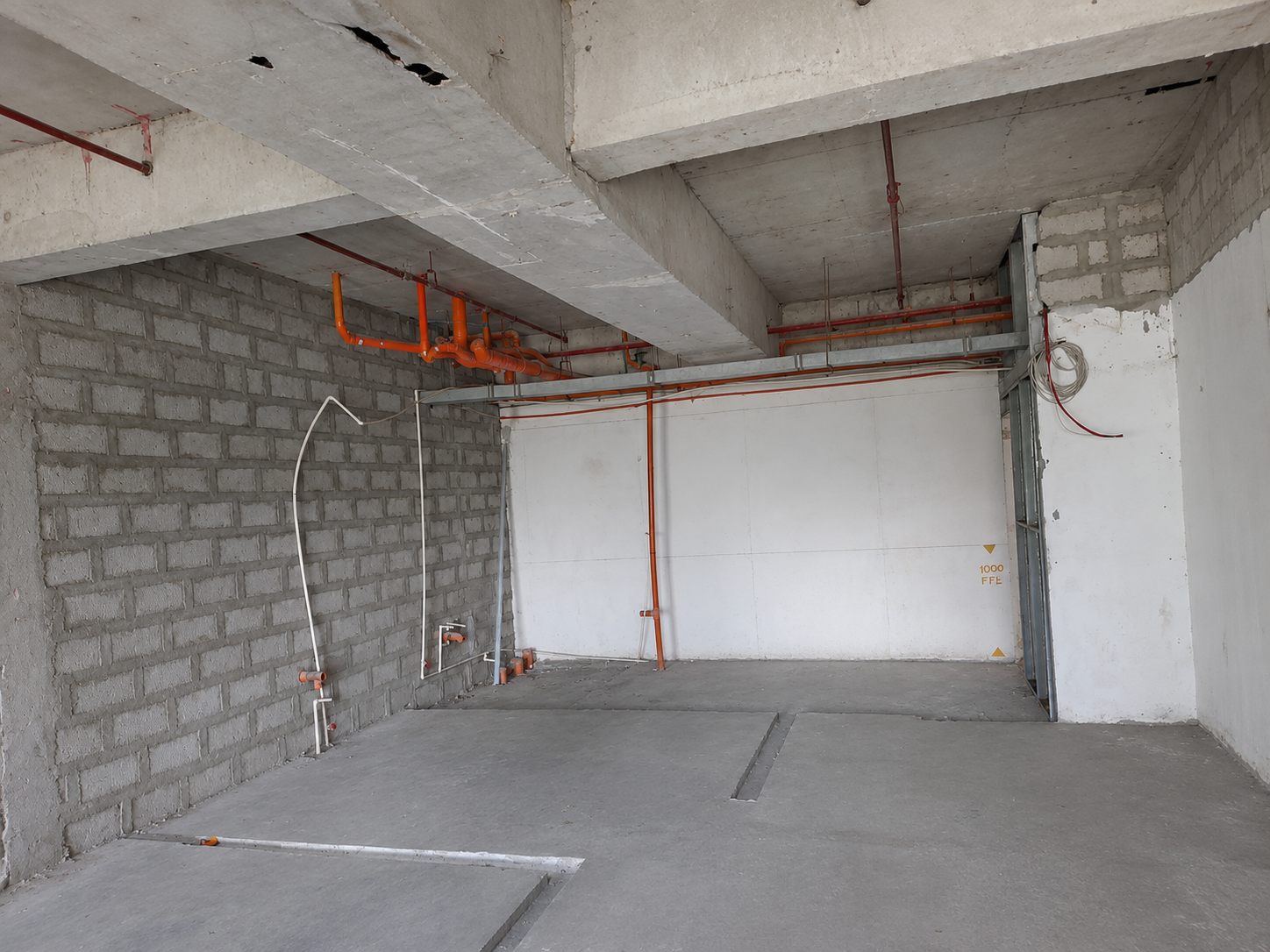 Under-construction room with orange plumbing pipes along the ceiling, concrete beams, and exposed wiring on the walls.
