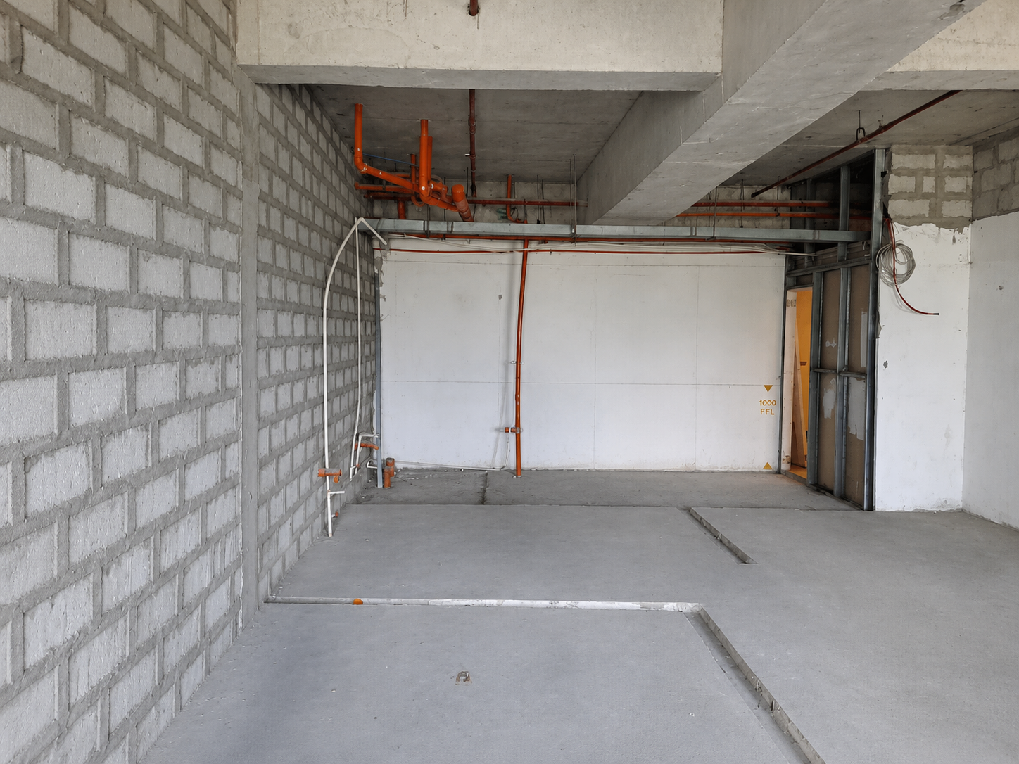 Construction site interior with exposed orange pipes along the ceiling and unfinished concrete walls and floor.