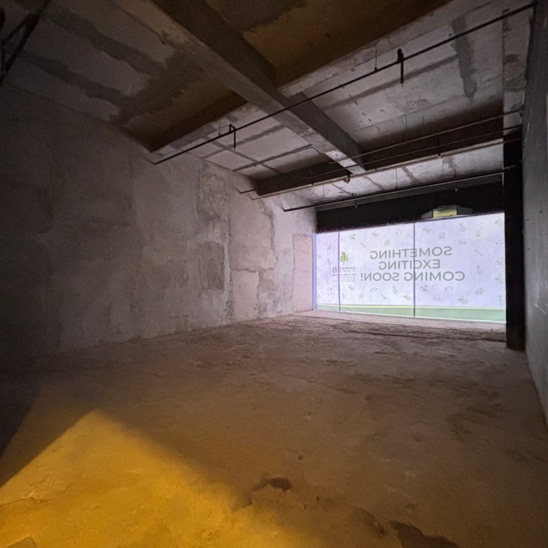 Dark unfinished under-construction hallway with concrete walls, exposed ceiling beams, and hanging cables receding toward a lit opening at the end.