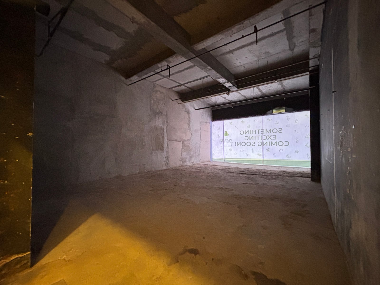 Unit 2-107_1 Dark unfinished under-construction hallway with concrete walls, exposed ceiling beams, and hanging cables receding toward a lit opening at the end.