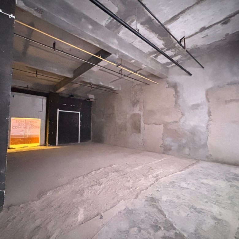 Industrial concrete interior with exposed ceiling pipes and a doorway, orange sign reading 'Something exciting coming soon!' nearby