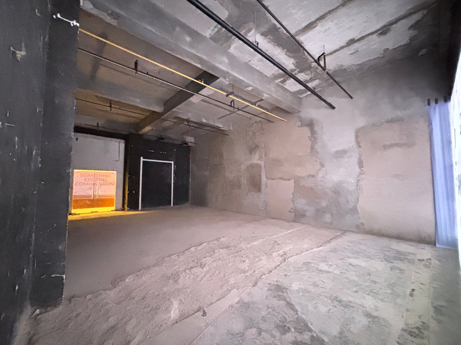 Unit 2-107_4 Industrial concrete interior with exposed ceiling pipes and a doorway, orange sign reading 'Something exciting coming soon!' nearby