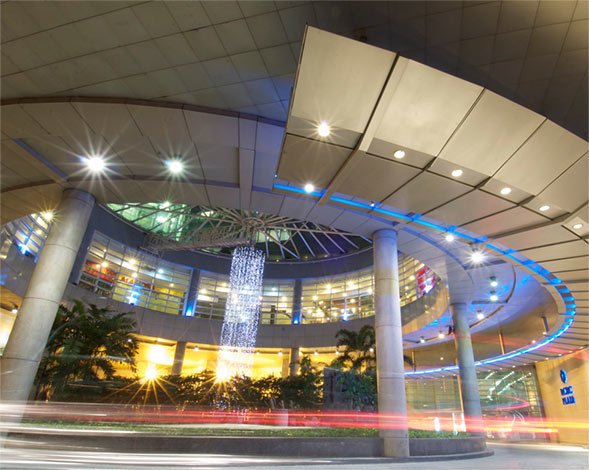 Exterior view of a circular modern building with blue neon rings, a tall illuminated sculpture in the center, and light trails from cars.