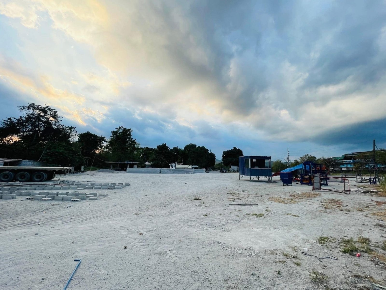 Open construction yard with scattered concrete blocks, trucks, and blue containers under a cloudy sky.