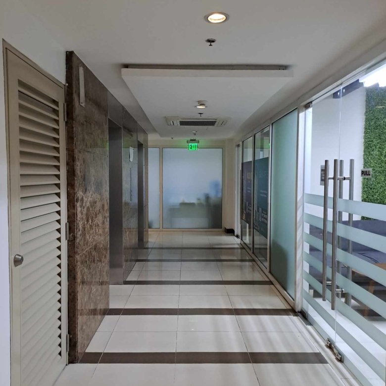 Office hallway with glass doors on the right, white tiled floor, marble wall accents, and a lit green exit sign in the distance.