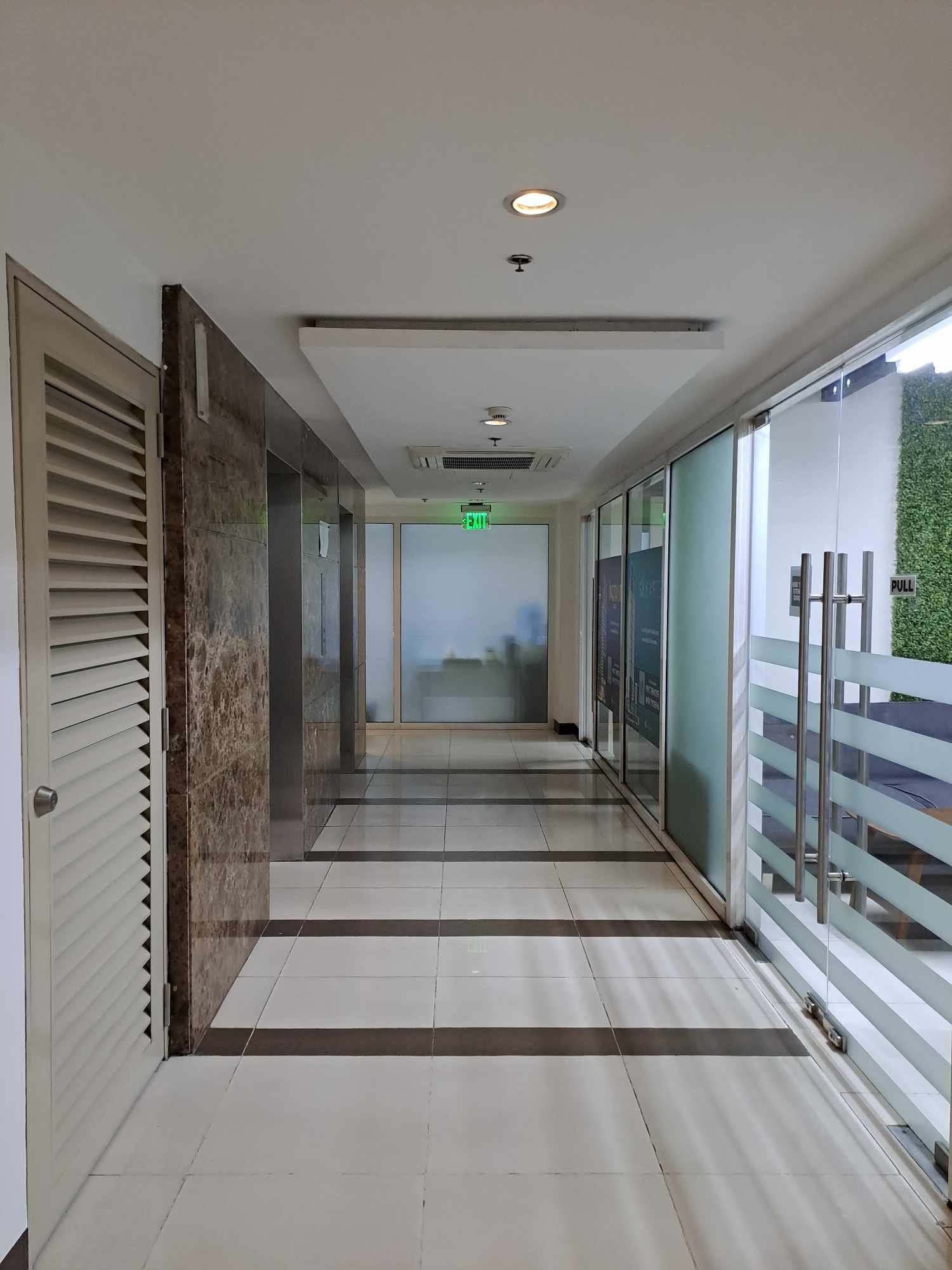 Office hallway with glass doors on the right, white tiled floor, marble wall accents, and a lit green exit sign in the distance.