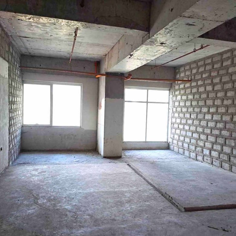 Empty unfinished room with concrete floor, brick walls, and two large windows; exposed piping and ceiling beams, under construction.