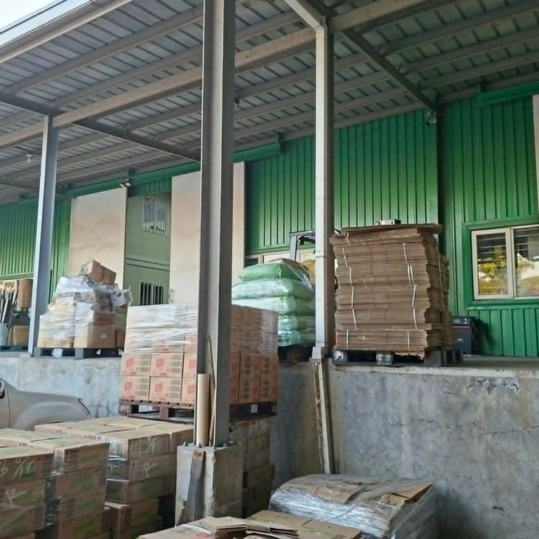 Industrial loading area with stacked cardboard boxes and pallets under a metal roof; a white car is parked to the left.
