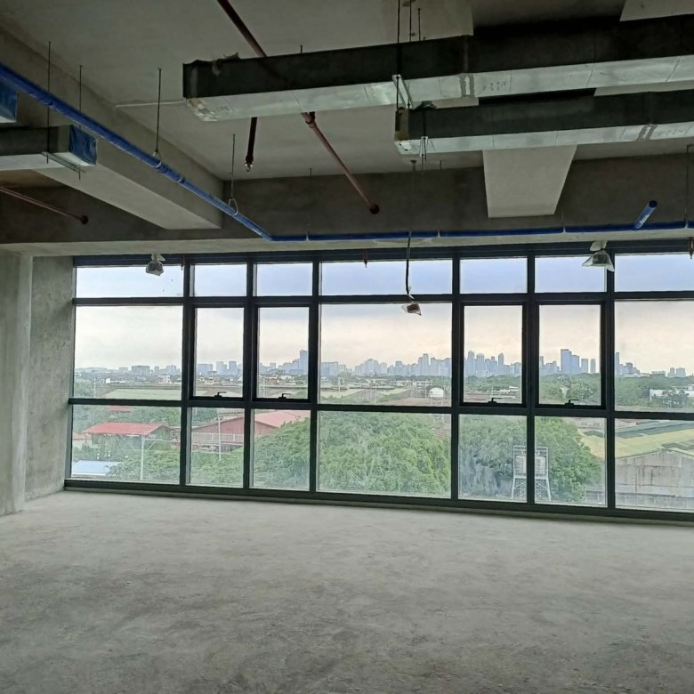 Empty unfinished office space with exposed concrete, hanging pipes, and large wall-to-wall windows offering a city skyline view in the distance.