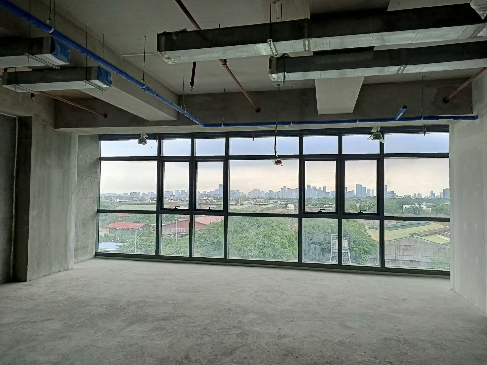 Empty unfinished office space with exposed concrete, hanging pipes, and large wall-to-wall windows offering a city skyline view in the distance.