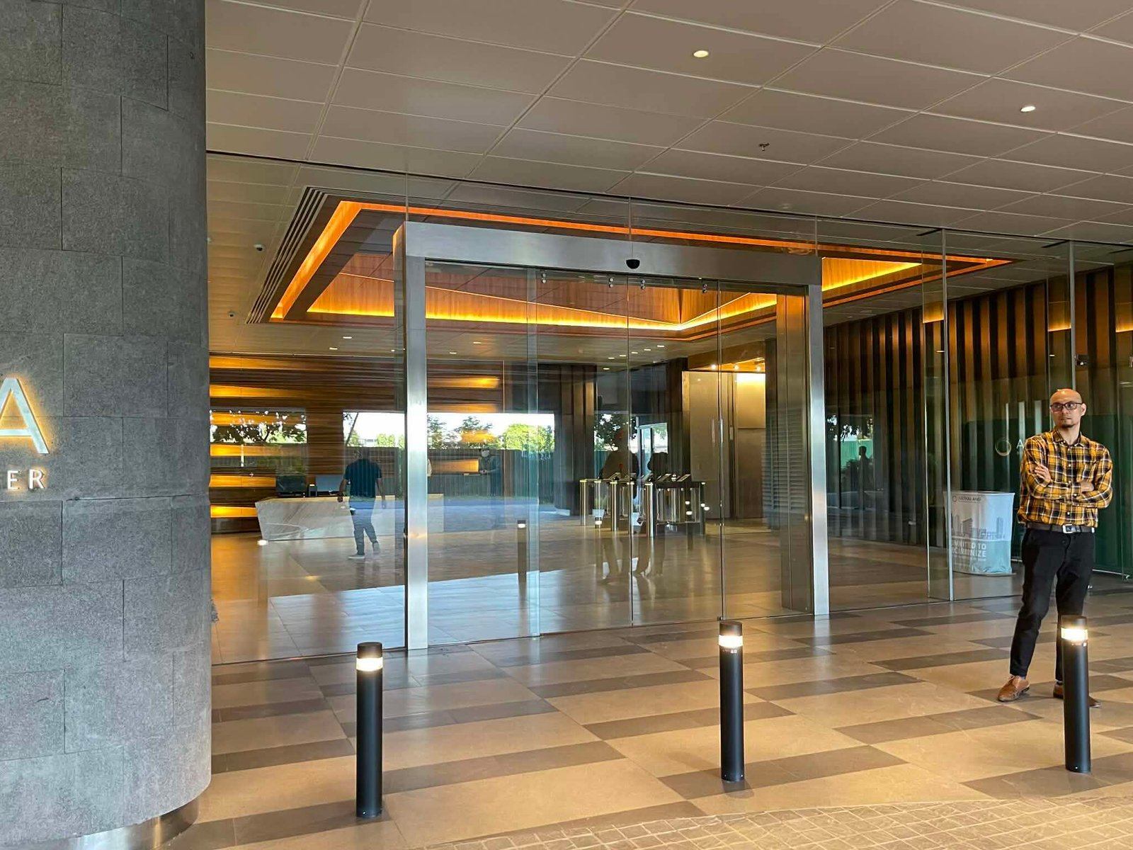 Modern office lobby with glass revolving doors, warm orange backlighting, and a man in a plaid shirt standing to the right near the entrance.