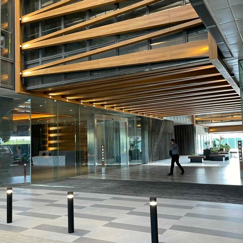Modern office lobby with glass walls, wood slat ceiling, and a person walking across a tiled floor.