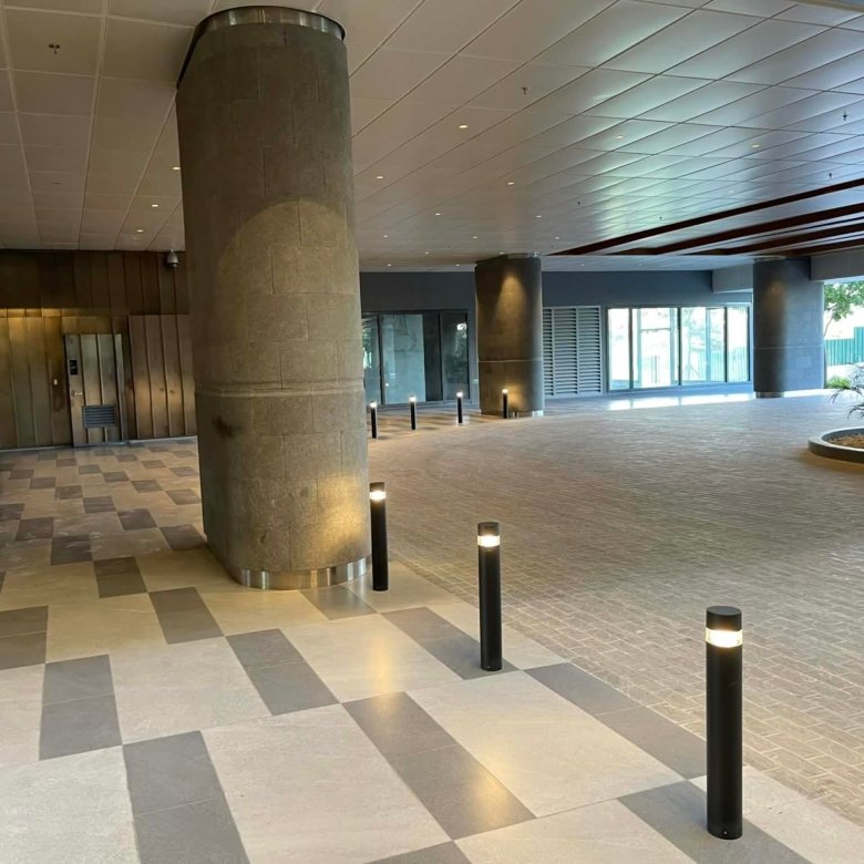 Indoor hotel or office lobby with large stone columns, glass doors, and a checkered tile floor; soft ambient lighting and short bollard lights present.