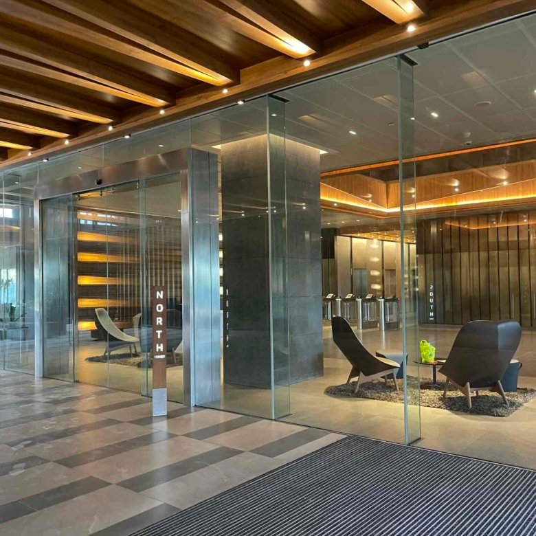Modern office lobby with glass walls, wooden ceiling beams, and lounge chairs set on a tiled floor.