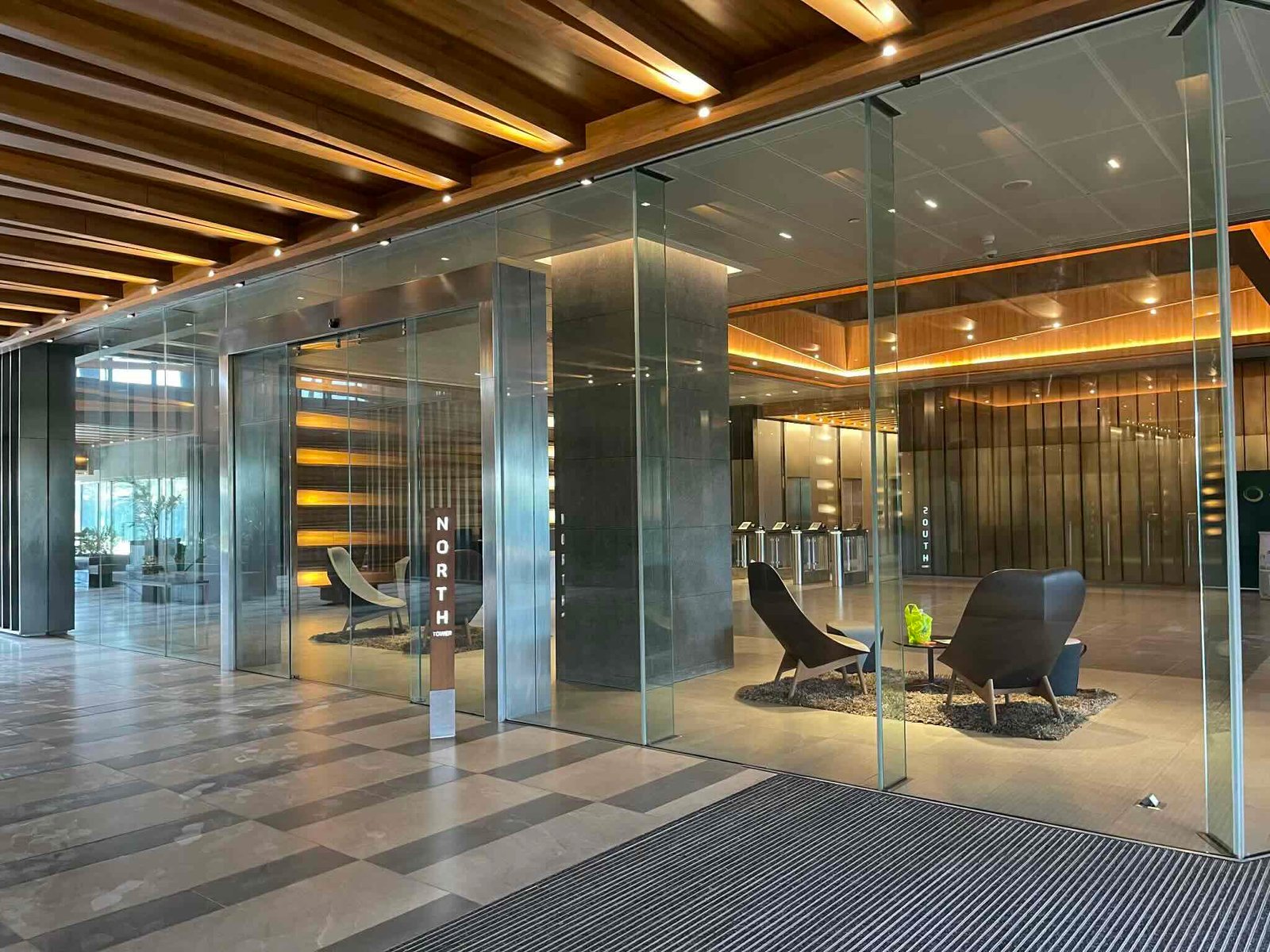 Modern office lobby with glass walls, wooden ceiling beams, and lounge chairs set on a tiled floor.