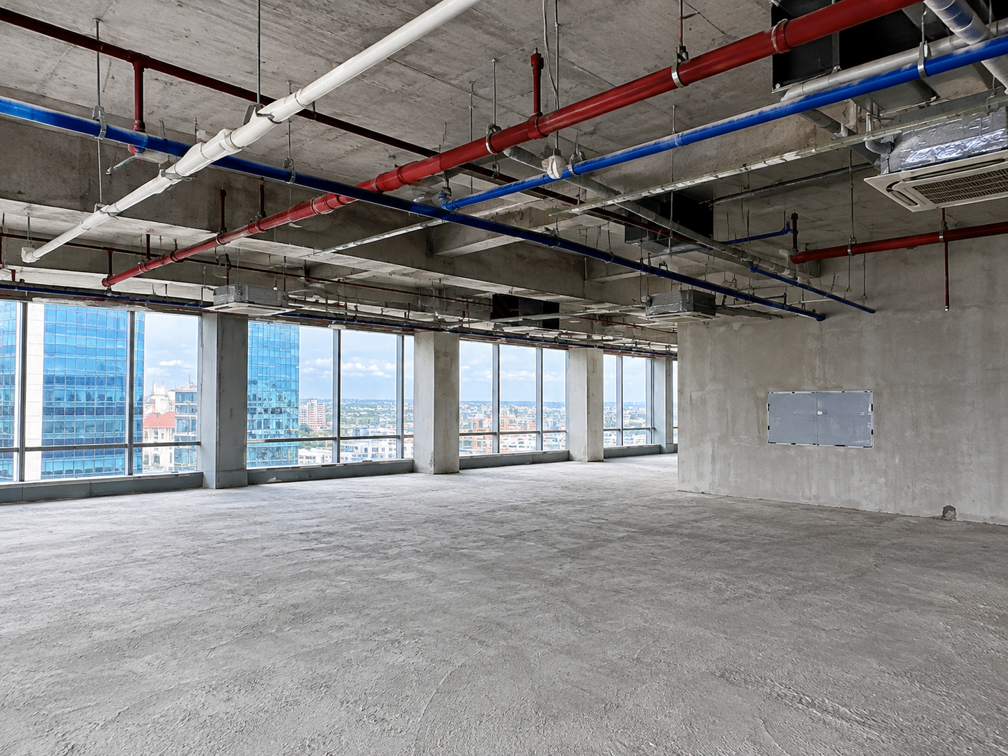 Empty unfinished office space with exposed plumbing and large city-view windows.