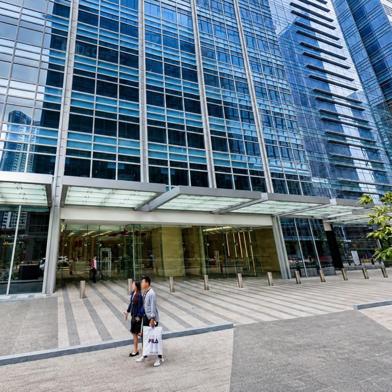 Two pedestrians walk along the entrance of a tall glass office building with a glass canopy, one carrying a white FILA shopping bag there.
