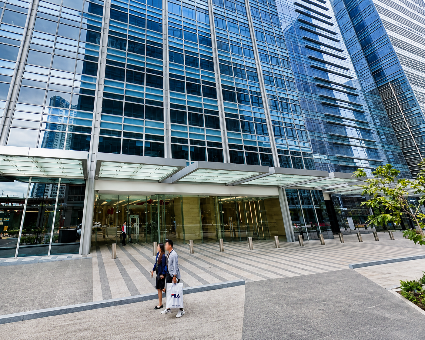 Two pedestrians walk along the entrance of a tall glass office building with a glass canopy, one carrying a white FILA shopping bag there.