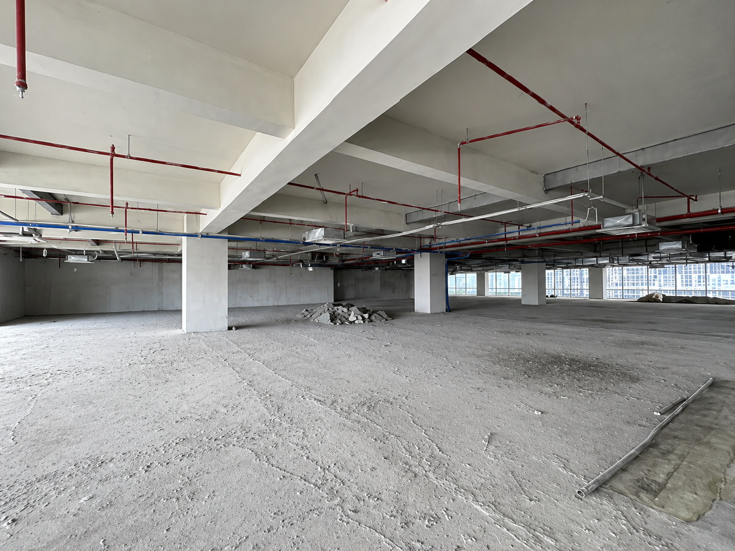 Open unfinished interior with concrete columns, red and blue pipes, ducts, and a pile of rubble on a dusty floor.
