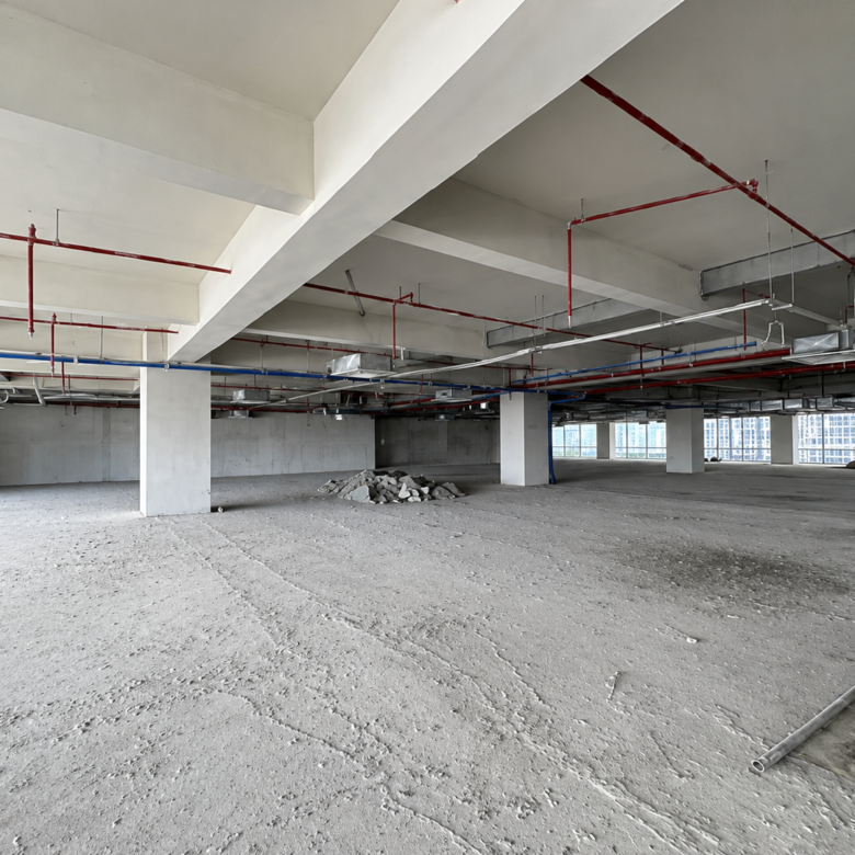 Open unfinished interior space with concrete floors, supporting columns, and exposed red and blue overhead pipes and ducts, under construction.