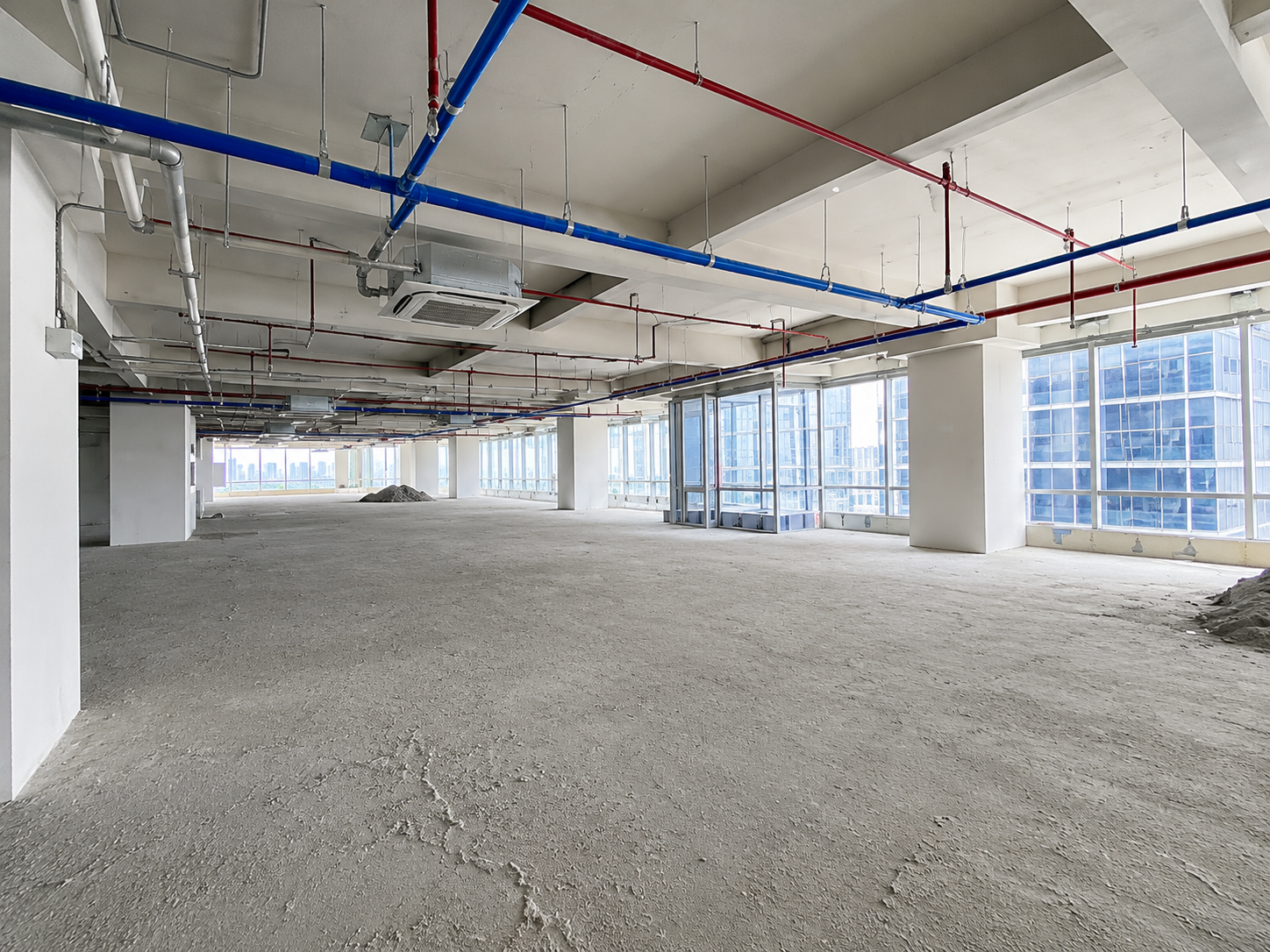 Unfinished open-plan office space with exposed red and blue pipes, concrete floor, and large windows showing a city skyline.
