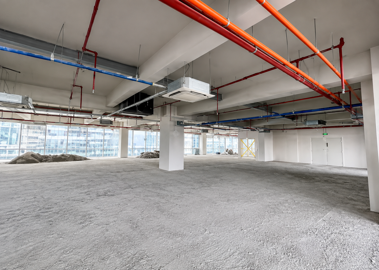 Open, under-construction office space with exposed red and blue pipes, concrete floor, and dirt piles by large windows.