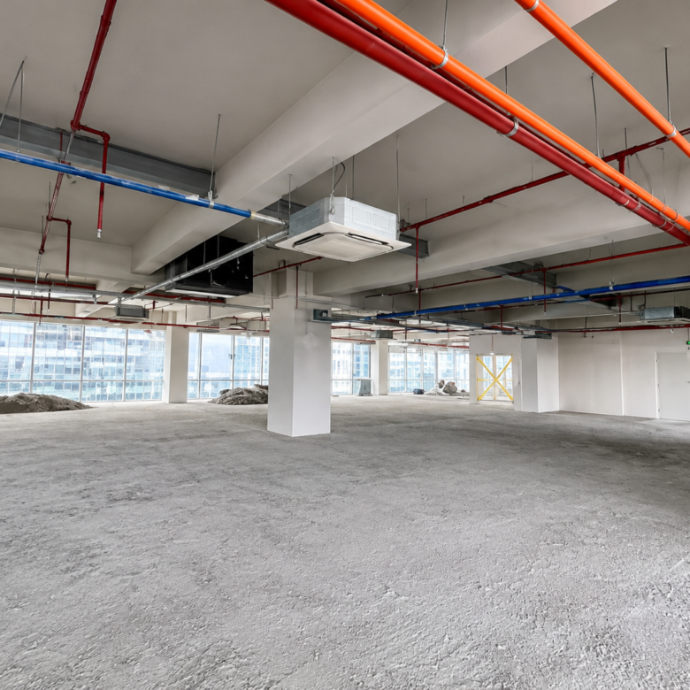 Open, unfinished interior of a high-rise with exposed red, orange, and blue pipes running along the ceiling and large windows at the far wall (construction site).Chipped concrete floor and a few dirt piles visible near the windows, indicating ongoing construction.