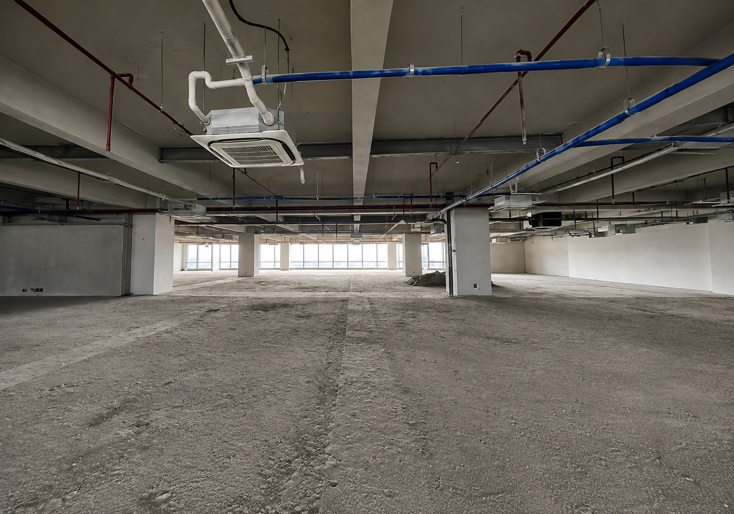 Empty unfinished basement space with concrete floor, large support columns, and exposed blue and white pipes overhead in a construction site. It appears to be an open, under-construction level with natural light from distant windows.