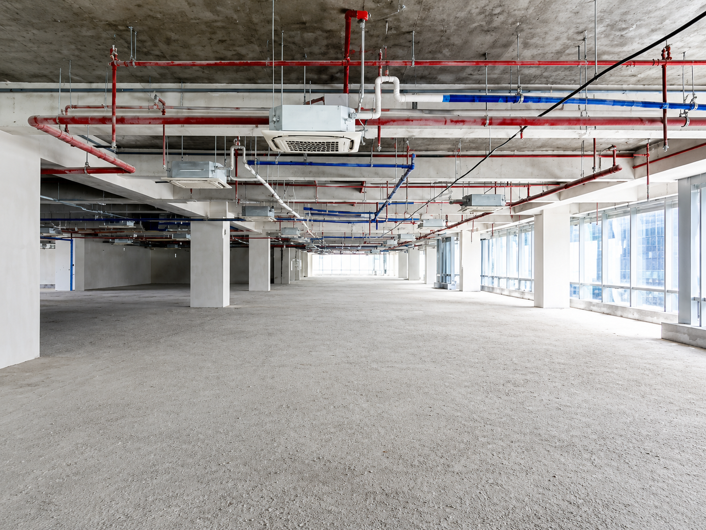 Empty unfinished commercial space with exposed red and blue ceiling pipes and large windows on the rightside wall.