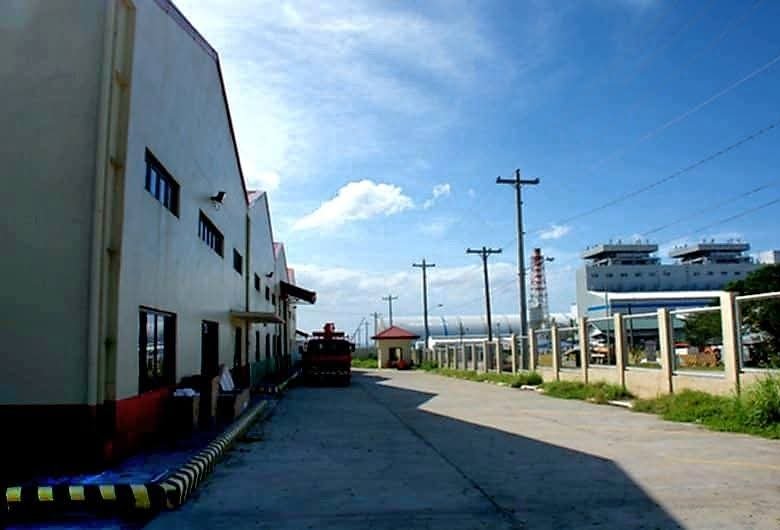 Industrial waterfront scene: a long white factory on the left, power poles along a paved road, and ships in the harbor on the right.