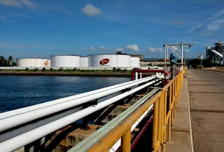 Industrial waterfront with large white storage tanks along the water and a network of yellow and white pipes on a railing, under a blue sky.
