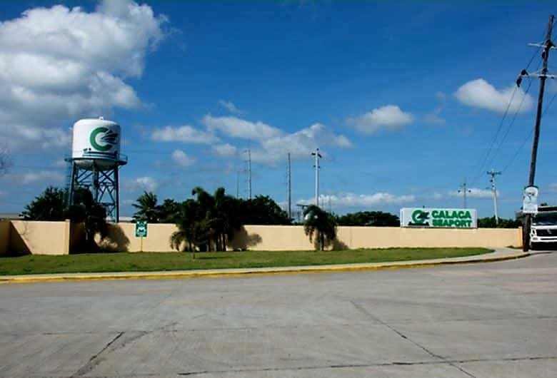 Entrance to Calaca Port: beige perimeter wall, large water tower with logo, palm trees, and a truck on the road under a blue sky.