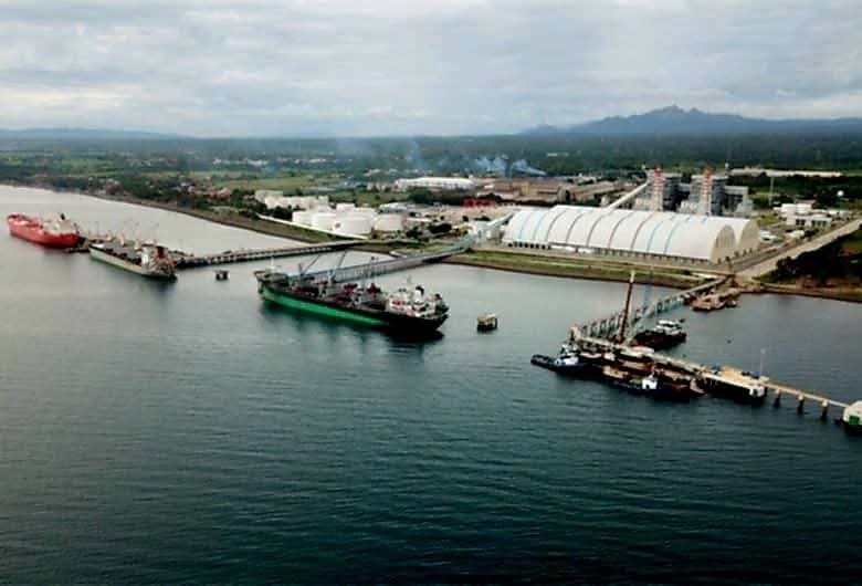 Aerial view of an industrial port with cranes, ships, and a long white curved-roof building along the water.