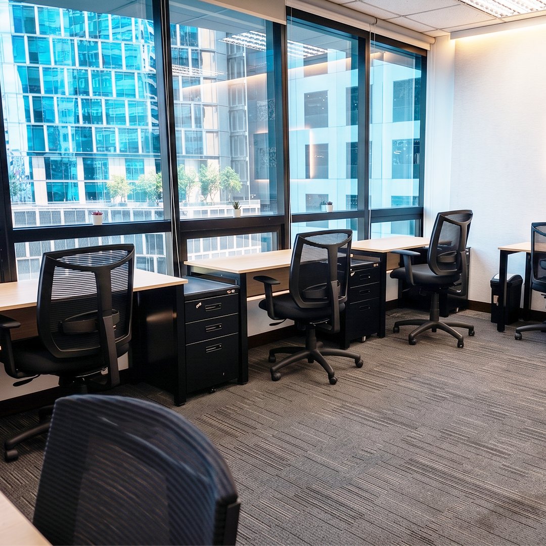 Row of workstations by large glass windows overlooking a city, with black mesh chairs and filing cabinets.