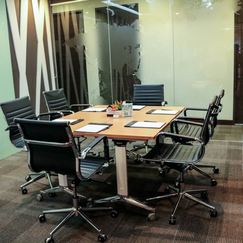Glass-walled conference room with a long wooden table, black rolling chairs, and papers in front of each seat.