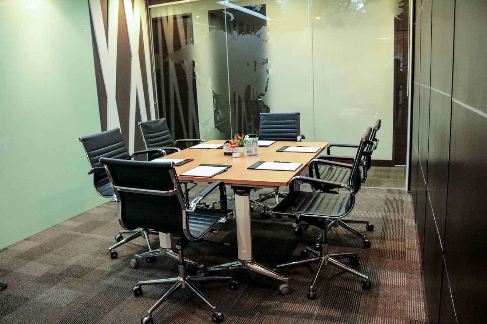 Glass-walled conference room with a long wooden table, black rolling chairs, and papers in front of each seat.