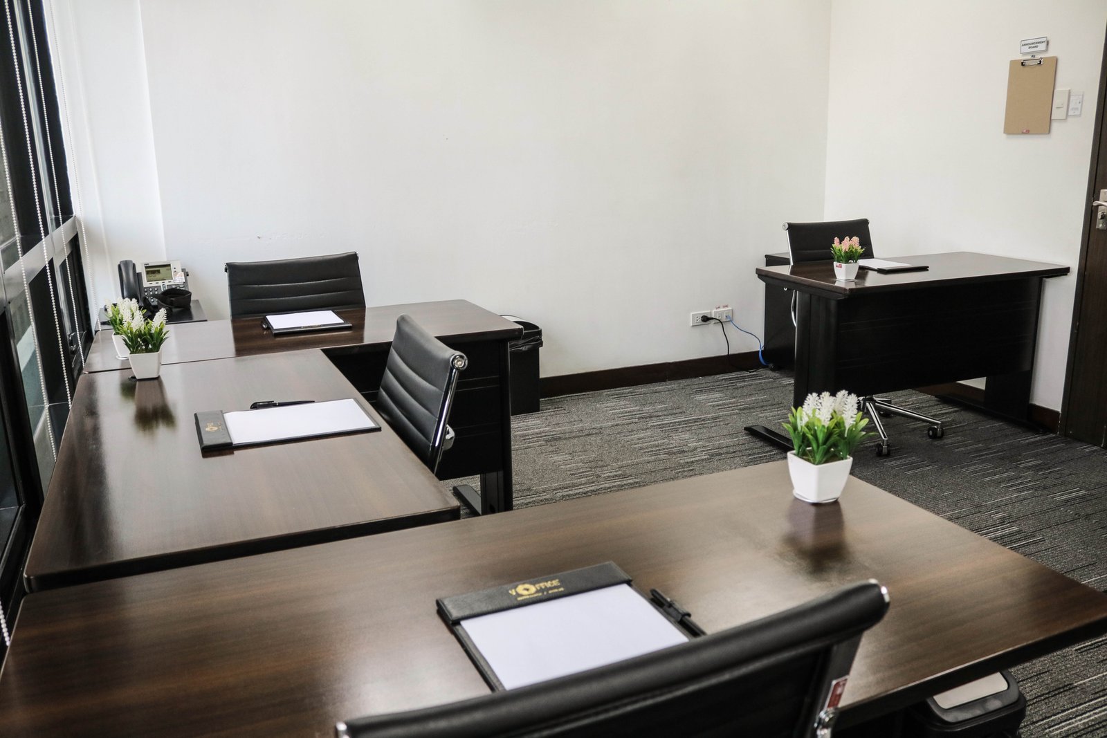 Empty office with dark wood desks, black chairs, and small white pots with green plants on each desk.",