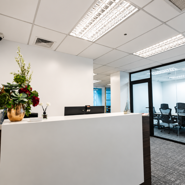 Reception area with a white front desk and a copper vase of red and green flowers; glass-walled meeting room visible to the right.