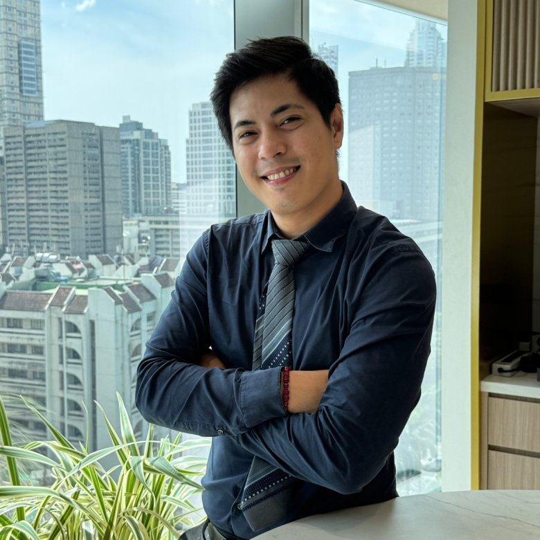 Smiling man in a navy shirt and striped tie stands with arms crossed near a large window overlooking a city skyline.